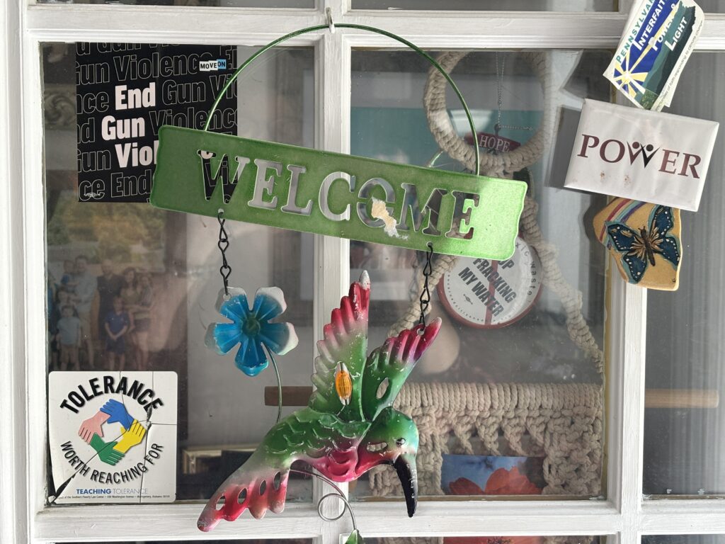 A green metal Welcome sign with a hummingbird and flower hanging in a window decorated with various political stickers.