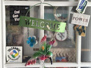 A green metal Welcome sign with a hummingbird and flower hanging in a window decorated with various political stickers.