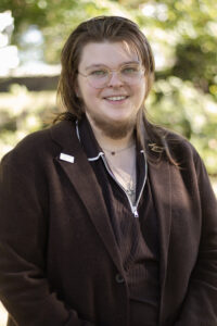 Portrait of a person with shoulder-length brown hair and glasses, wearing a brown blazer.