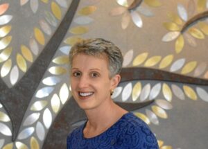 A woman with short grey hair smiling in front of a decorative wall with metal leaf shapes.