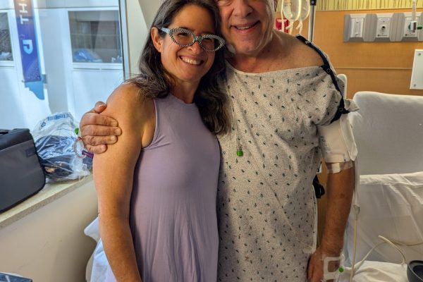 Smiling woman hugs man in hospital gown with medical equipment, both standing in a bright hospital room.