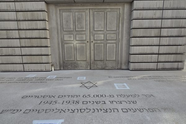 A stone memorial with engraved Hebrew text stands in front of a gray building under a partly cloudy sky.