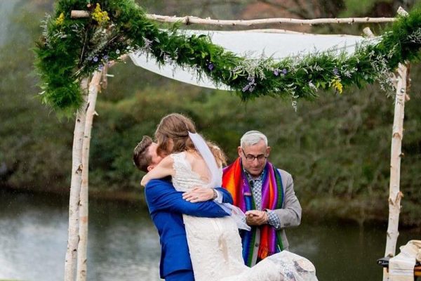 queer couple - one in a white bridal gown and one in a blue suit - embracing under a chuppah with a rabbi wearing a rainbow tallit