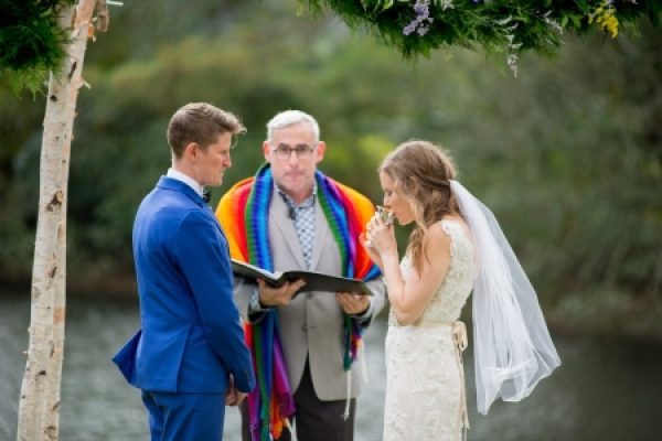 A couple stands facing each other at an outdoor wedding ceremony near a river, with an officiant in colorful attire.