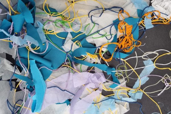 Colorful fabric scraps and threads are scattered on a dark surface, creating an abstract, tangled composition.