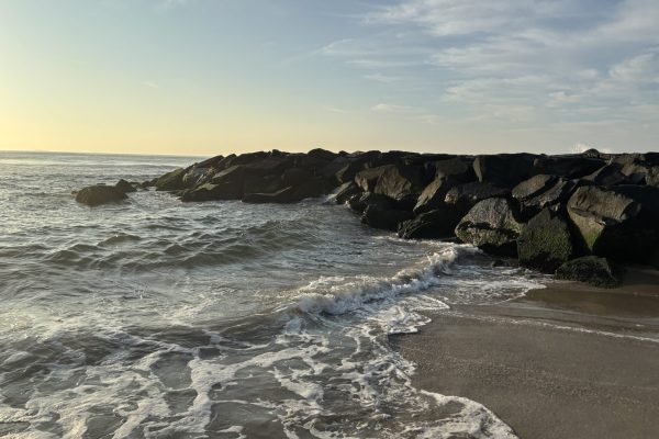 Waves gently washing onto a sandy beach with a rocky jetty under a partly cloudy sky at sunset.