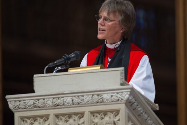 Rev. Budde in religious attire speaks at a podium with ornate carvings and a microphone.