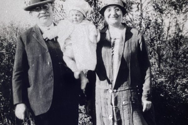 Black and white photo of two adults and a baby standing outdoors, all dressed in vintage clothing.