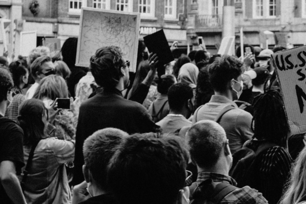 Crowd at a protest, holding signs. Urban street setting with old buildings in the background.