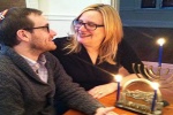 A man and woman smiling at each other beside a lit menorah on a table.
