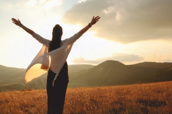 Person with outstretched arms in a field at sunset, facing distant hills and a cloudy sky.