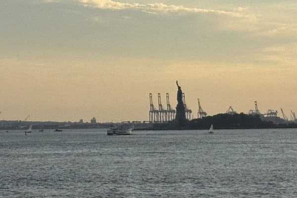 The Statue of Liberty and cranes are seen at sunset across the water, with boats and a cloudy sky.