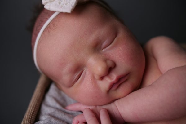 Sleeping newborn wearing a headband with a bow, resting peacefully against a soft blanket in a dark setting.