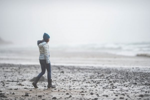 Person in a blue beanie and winter coat walking along a foggy, rocky beach.