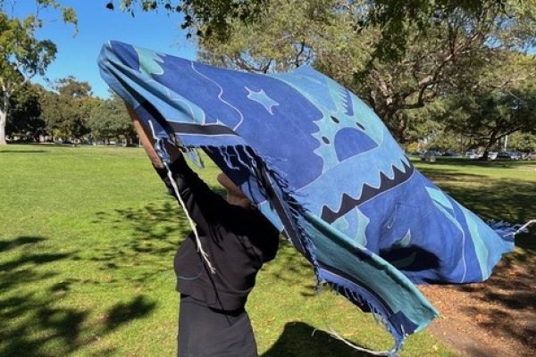 Person outdoors holding up a large blue patterned blanket as it billows in the wind under trees.