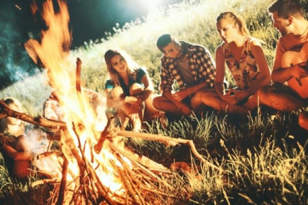 Group of people sitting around a campfire in the grass, one person playing a guitar.