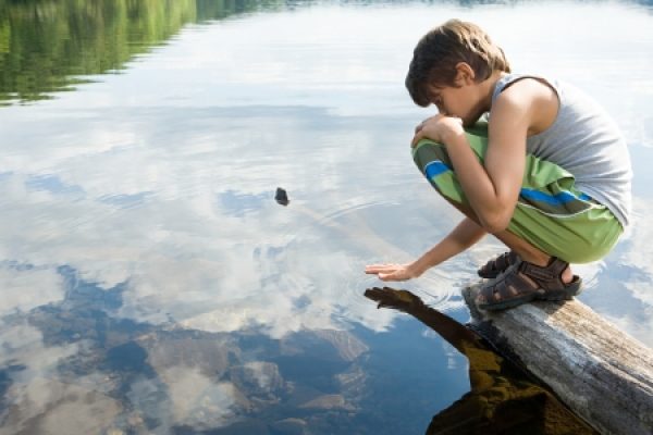 A child crouches on a wooden log, touching the surface of a calm lake with stones visible underwater.