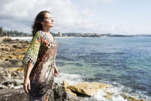 Woman in colorful dress stands on rocky shore, eyes closed, enjoying the breeze by the sea.