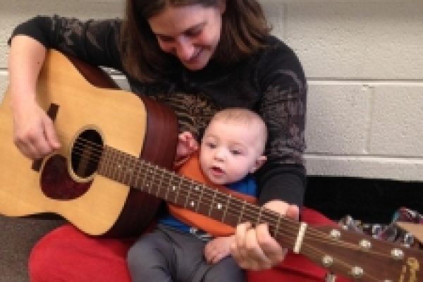 Woman holding a baby and playing a guitar, both seated on the floor, smiling.