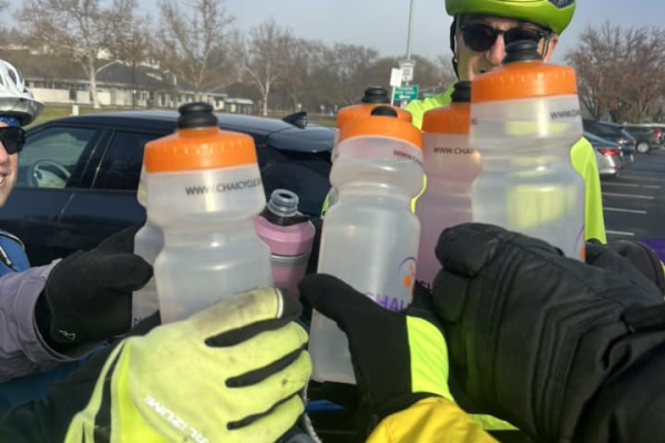 Cyclists wearing helmets toast with water bottles in a parking lot on a cloudy day.