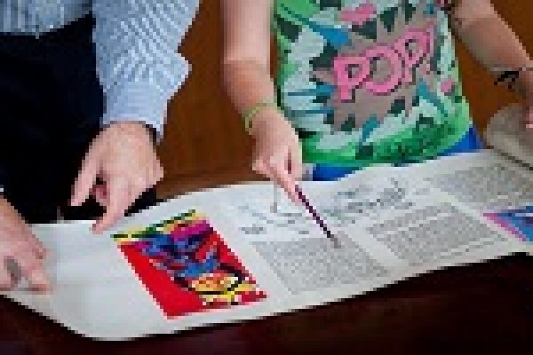 Two people examine a large megillah scroll with colorful illustrations and Hebrew text, pointing at different sections.