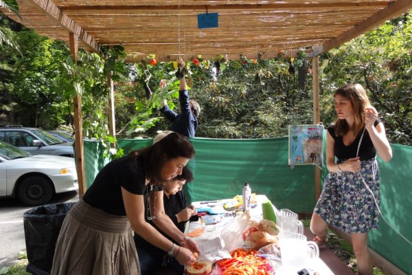 People preparing food under a leafy canopy, with snacks on a table and a person hanging decorations.