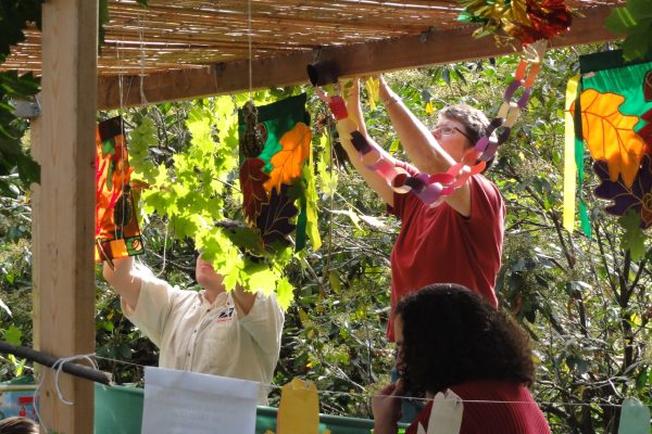 People decorating a sukkah with foliage and colorful paper chains under a wooden roof.
