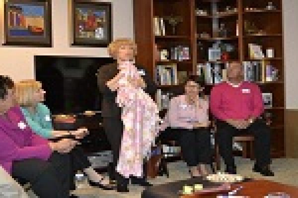 A woman stands holding a baby in a cozy living room with several seated people watching attentively.