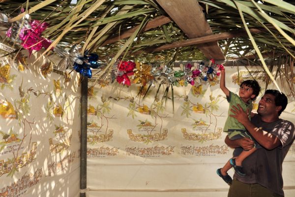 A father lifts his child to reach colorful decorations inside a bamboo-roofed sukkah.