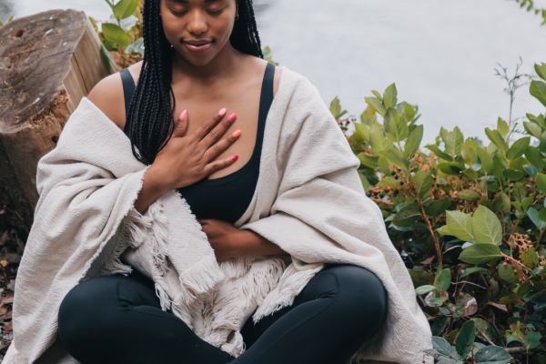 Woman sitting cross-legged by water, eyes closed, hand on chest, wrapped in a cozy blanket outdoors.