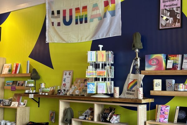 Colorful shop display with pride-themed items, books, and a HUMAN banner on a yellow and blue wall.