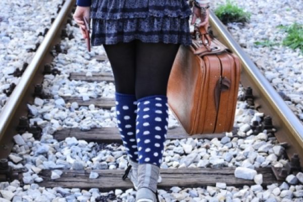 Person walking on train tracks with a brown suitcase, wearing a frilly skirt, polka-dot socks, and boots.
