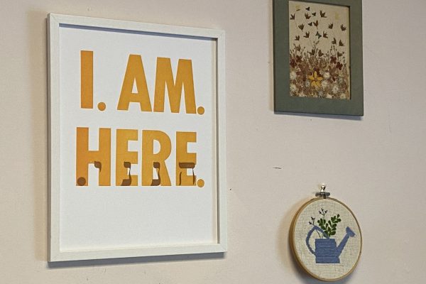Three framed artworks on a wall, one reads “I AM. HERE.” in bold yellow letters.