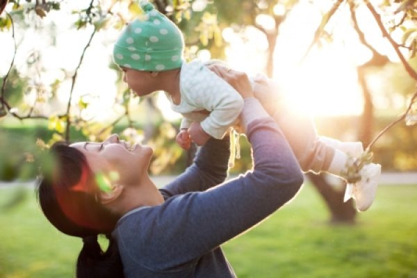 Woman joyfully lifts a smiling baby in nature, with sunlight streaming through trees in the background.