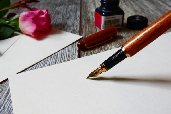 A fountain pen on paper with a rose, an ink bottle, and cap on a wooden surface.