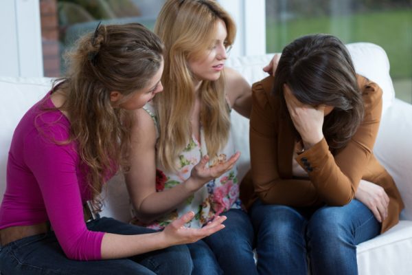 Three women sitting on a couch; two are comforting the third person, who is covering her face with her hand.