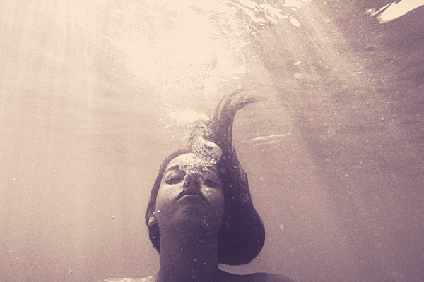 Woman underwater blowing bubbles, with light rays filtering through the water from above.
