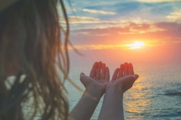 Person with open hands silhouetted against an ocean sunset.
