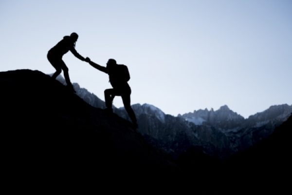 Silhouettes of two hikers helping each other on a mountain with a clear sky and rugged peaks in the background.