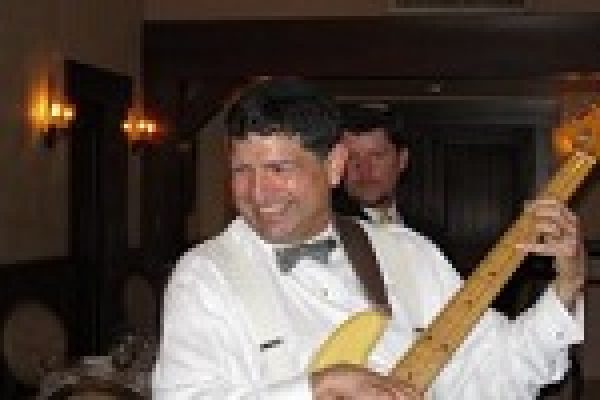 Man in a white suit playing a guitar at an indoor event, smiling with people in the background.