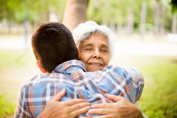 An older woman and a younger person embrace warmly in a park setting.