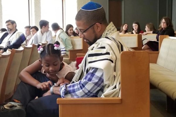 black male Jewish person wearing a tallit and blue kippah, sitting in the pews of a synagogue, as a little black girl in a pink dress sits on his lap. He is wearing glasses and looking down at her. Other congregants are in the background.