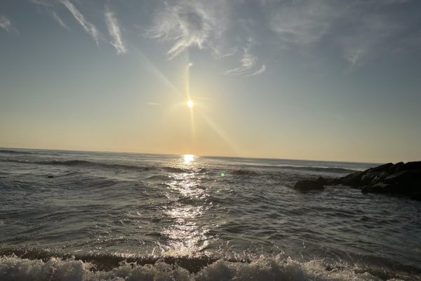 Sunset over the ocean, waves gently crashing near rocks, and clouds scattered across the sky.