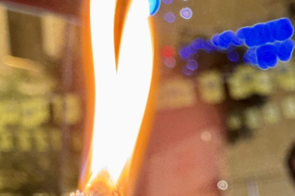Close-up of a blue lighter with two flames, blurred city lights and a poster in the background.