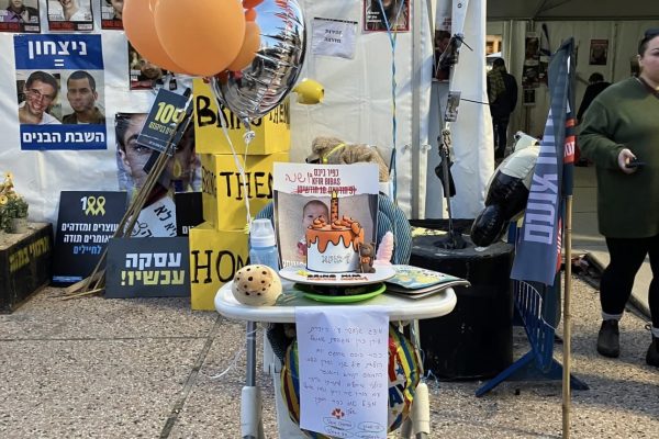 A highchair with orange balloons, protest signs, and a sign in Hebrew at an outdoor event with people nearby.