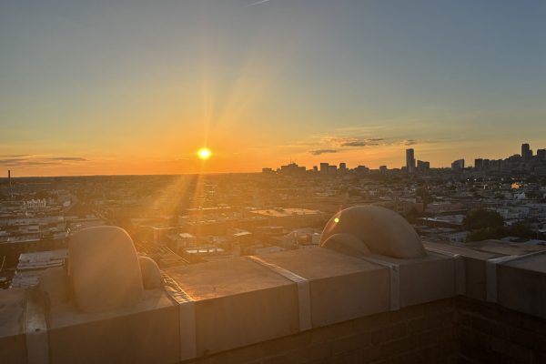 Bright orange sunset over a city skyline, viewed from a rooftop with stone ledges in the foreground.