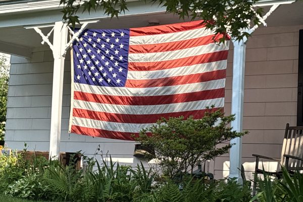Large American flag hangs vertically on the porch of a house, garden plants in front, rocking chair to right.