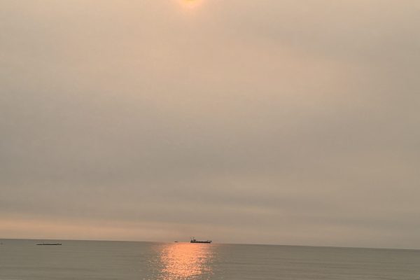 The sun glows orange over a calm ocean with a distant ship and shimmering reflection on the water.