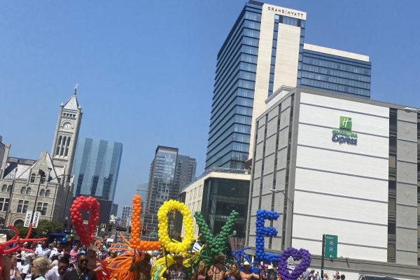 People marching in a Pride parade holding colorful balloon letters spelling LOVE on a sunny city street.