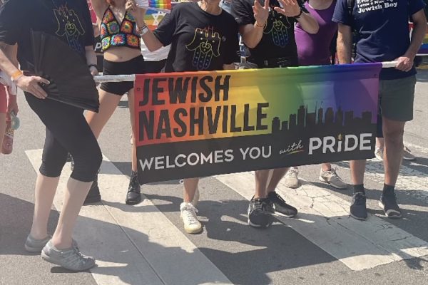 A group holds a Jewish Nashville Welcomes You with Pride banner at a Pride parade on a sunny day.